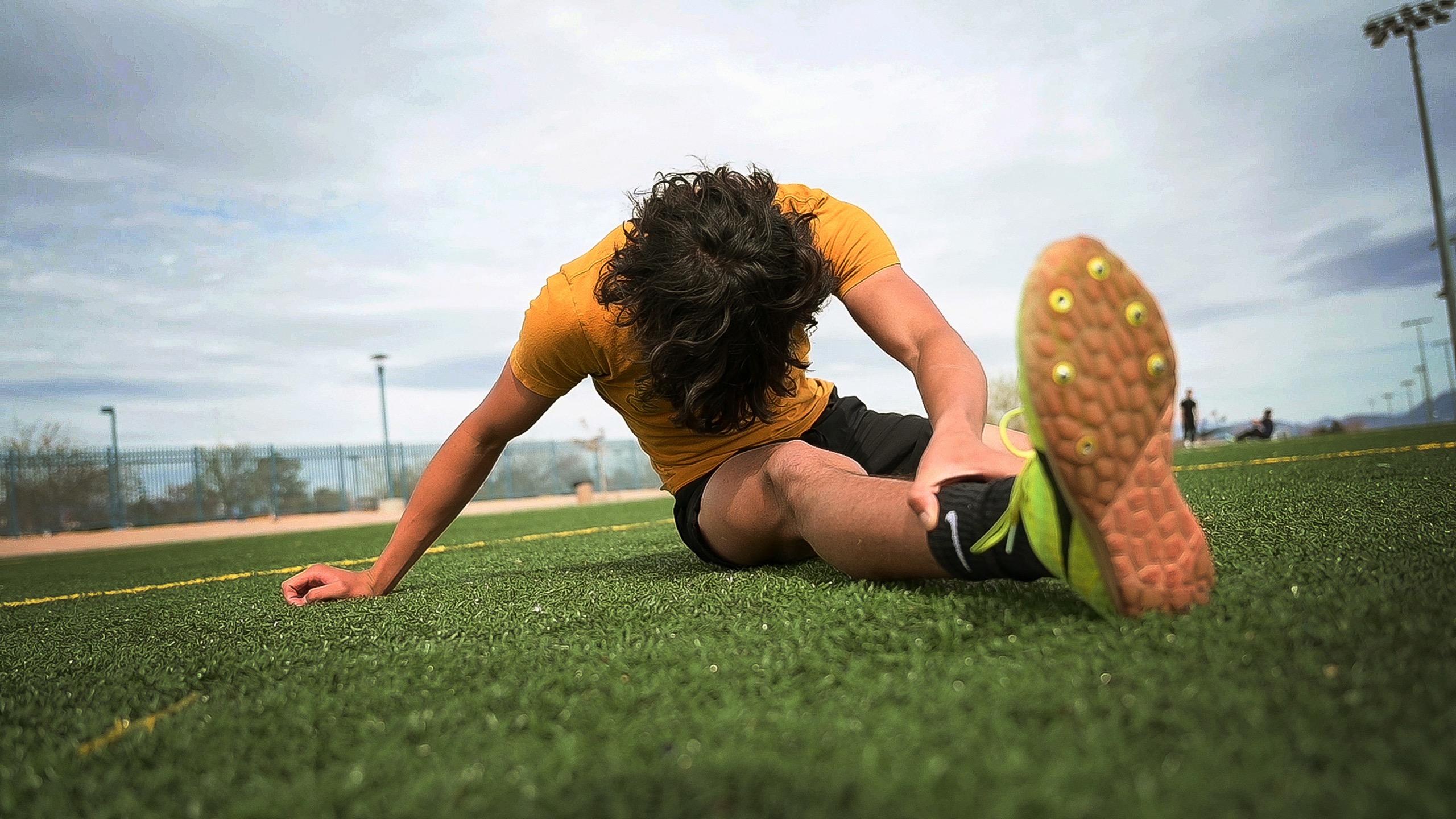 Track athlete in Nevada State colors performing a dynamic hamstring stretch on the turf, demonstrating the preparation and flexibility required for peak performance